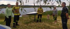 Wspólne patrolowanie akwenów. Na plaży stoi dwóch strażaków, policjant oraz dwie kobiety. W tle akwen, kajak. Z lewej strony widoczny jest fragment pomostu. Strażacy maja ubrane bluzy koloru piaskowego oraz czarne spodnie.