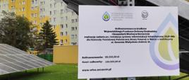 Na zdjęciu znajduje się tablica informująca o dofinansowaniu ze środków Wojewódzkiego Funduszu Ochrony Środowiska i Gospodarki Wodnej w kwocie 60 000 zł.
Całkowity koszt 120 000 zł. Tablicę zawieszono na metalowej konstrukcji, na której spoczywają ogniwa fotowoltaiczne.