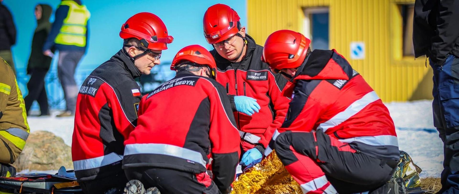 Zdjęcie przedstawia czterech ratowników medycznych w trakcie akcji ratunkowej, są to ćwiczenia w terenie górzystym lub pagórkowatym. Mężczyźni klęczą w kręgu na suchej trawie, pochylając się nad poszkodowaną osobą, która jest niemal całkowicie przykryta złotą folią izotermiczną (kocem NRC).