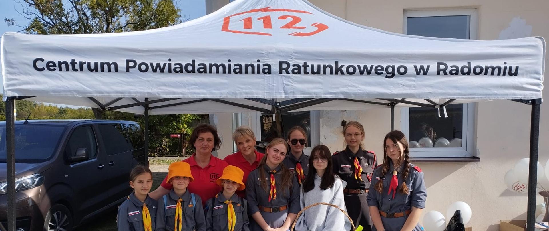 Dwie operatorki numeru 112 ubrane w czerwone koszulki oraz inni uczestnicy festynu pozują pod białym namiotem z napisem "112 Centrum Powiadamiania Ratunkowego w Radomiu" obok nich na trawniku leży kilka białych baloników. 