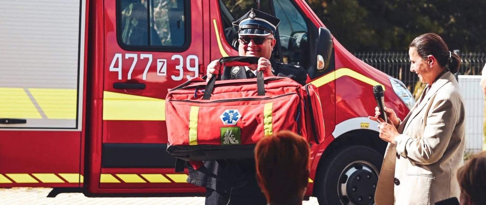 Na tle wozu bojowego straży pożarnej mężczyzna w stroju Ochotniczej straży Pożarnej podnosi prezentuje do zdjęcia czerwoną torbę z zestawem pierwszej pomocy przedlekarskiej z logo KRUS. Obok stoi kobieta w jasnej marynarce z mikrofonem w dłoni.
