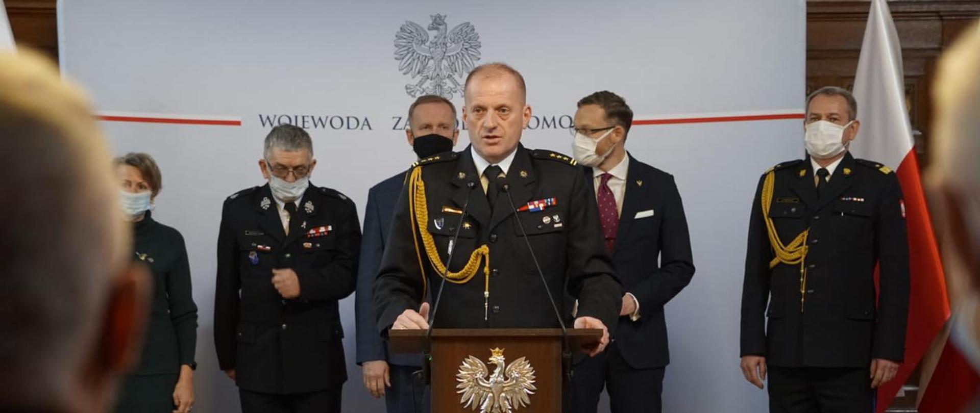 Przy mównicy st. bryg. Jarosław Tomczyk, za nim zaproszeni goście. W tle flagi Polski oraz baner wojewody zachodniopomorskiego.