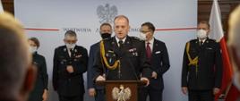 Przy mównicy st. bryg. Jarosław Tomczyk, za nim zaproszeni goście. W tle flagi Polski oraz baner wojewody zachodniopomorskiego.