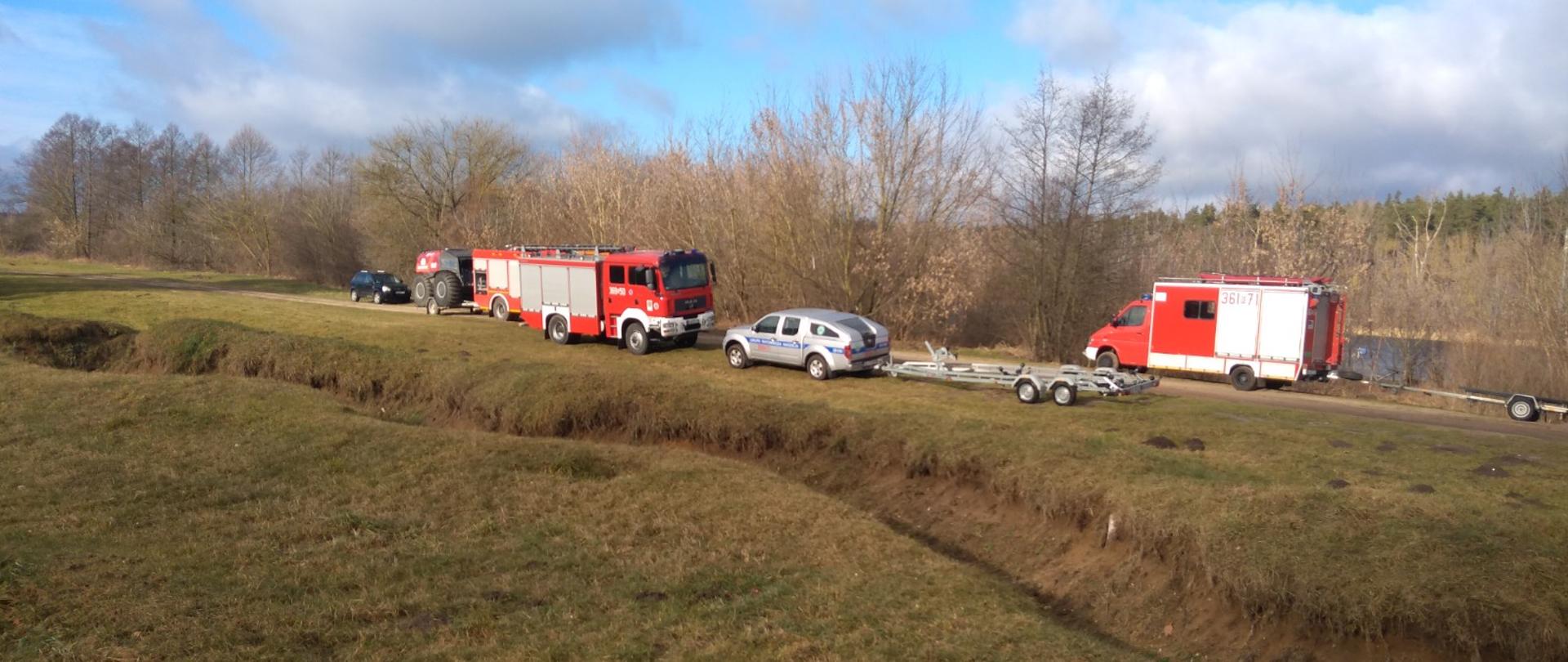 Ustawione samochody pożarnicze oraz lawety do przewodu łodzi na skraju rzeki Narew
