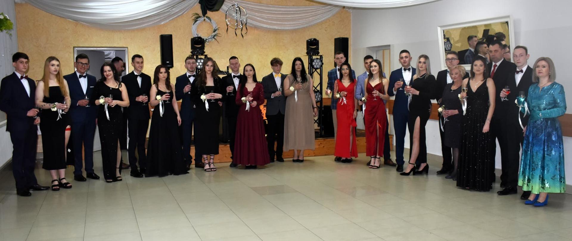 Grupa maturzystów i nauczycieli stoi w rzędzie na sali balowej, trzymając kieliszki podczas toastu. Sala udekorowana jest białymi i zielonymi materiałami pod sufitem.