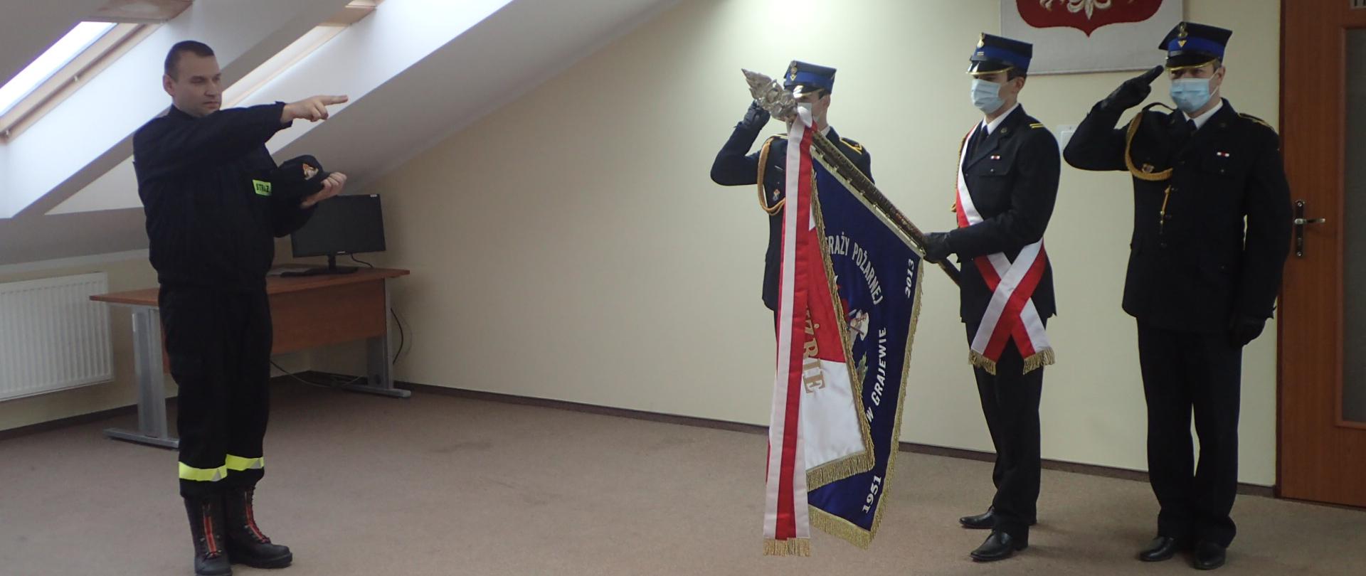 Na zdjęciu po lewej stronie stoi nowy funkcjonariusz, który składa ślubowanie na sztandar Komendy Powiatowej Państwowej Straży Pożarnej w Grajewie. Ubrany jest w ubranie koszarowe koloru czarnego, prawa ręka wyciągnięta wystawionymi dwoma palcami do ślubowania na sztandar. Po prawej poczet sztandarowy w pełnym umundurowaniu wyjściowym. Dowódca pocztu i asystujący salutują w trakcie roty ślubowania, za nimi ściana koloru jasnego, na której wisi biały orzeł na czerwonym tle.