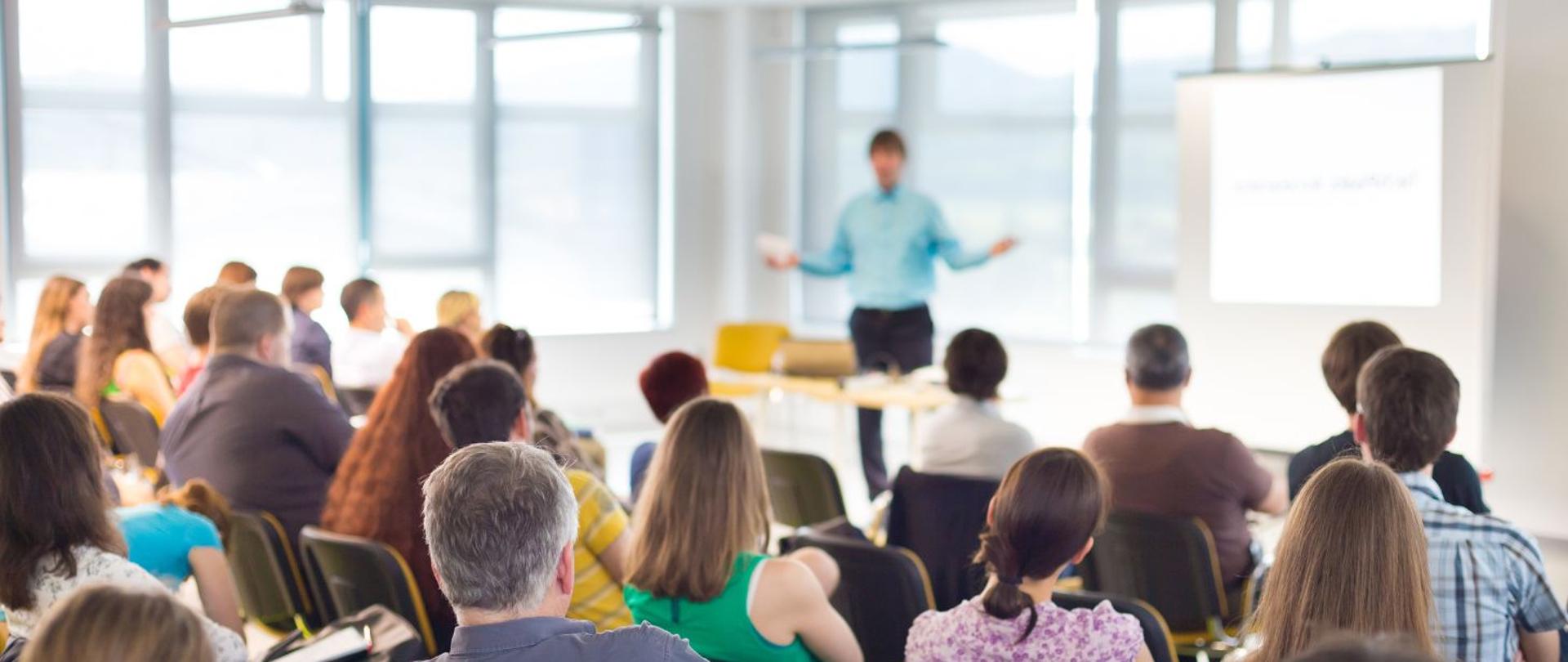 na zdjęciu widoczna jest grupa osób, siedząca w jednym pomieszczeniu. Na środku sali widać sylwetkę mężczyzny, który przemawia. 