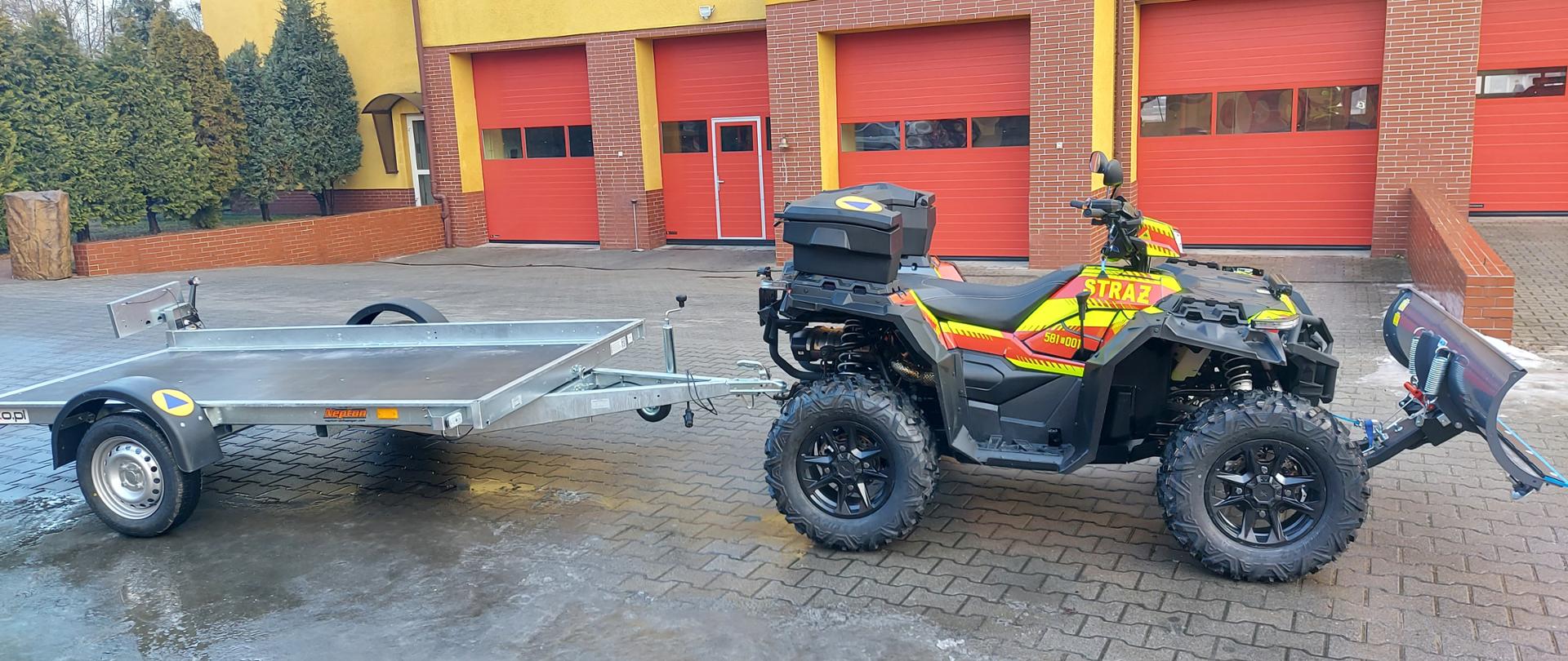 Zdjęcie przedstawia nowoczesny quad ratowniczy z przyczepą, zaparkowany na placu przed budynkiem straży pożarnej. Czarny quad marki Polaris z jaskrawymi, żółto-czerwonymi elementami odblaskowymi. Na boku pojazdu widnieje wyraźny napis „STRAŻ” oraz numery operacyjne. Pojazd jest wyposażony w czarny kufer wyprawowy zamontowany na tylnym bagażniku oraz pług do odśnieżania zamocowany z przodu. Do quada podpięta jest lekka, jednoosiowa przyczepa platformowa z niskimi burtami, służąca do transportu dodatkowego sprzętu lub osób poszkodowanych na specjalistycznych noszach. W tle znajduje się fragment budynku remizy strażackiej z trzema dużymi, czerwonymi bramami garażowymi. Plac wyłożony jest szarą kostką brukową, która jest miejscami mokra. Zdjęcie wykonane z boku, ukazujące pełną długość zestawu quad-przyczepa.