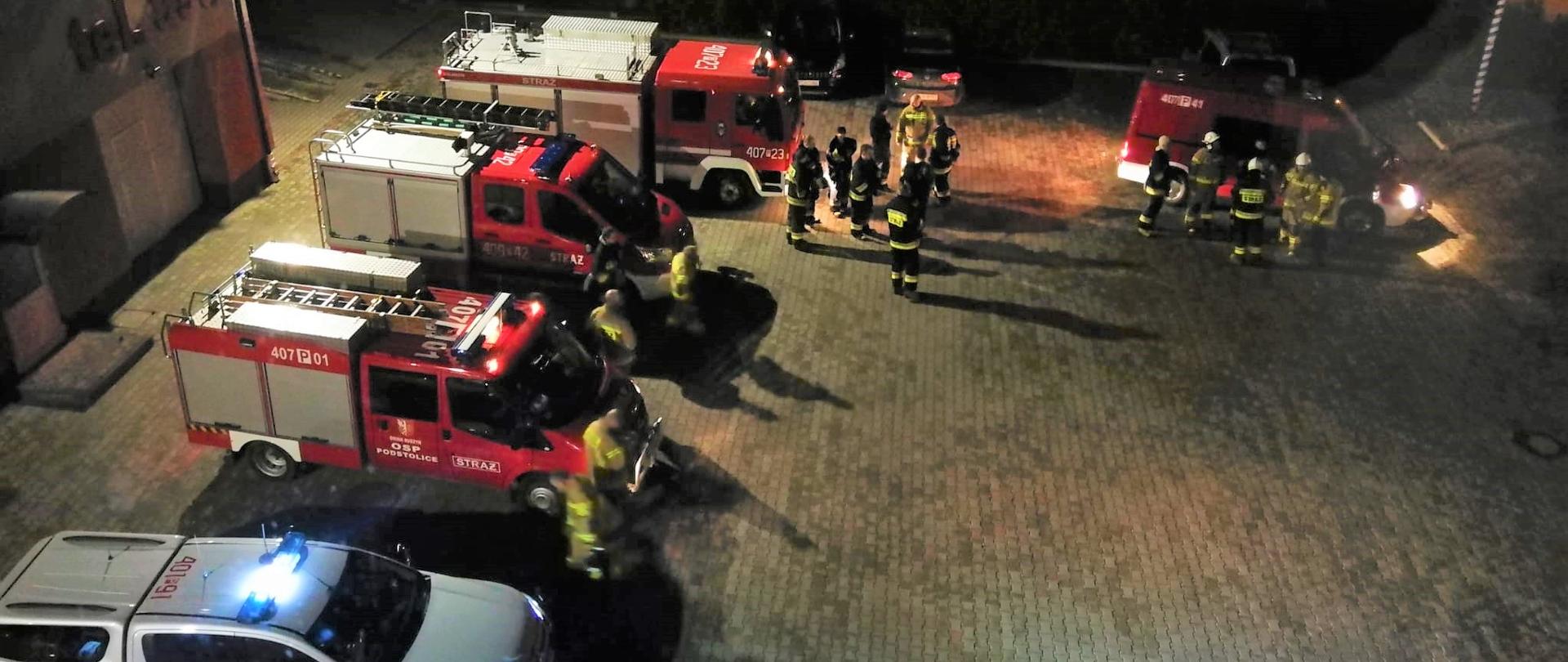 Zdjęcie przedstawia strażaków szykujących się do poszukiwań nocnych zaginionej osoby oraz samochody strażackie.
W tle plac Komendy oraz budynki.

