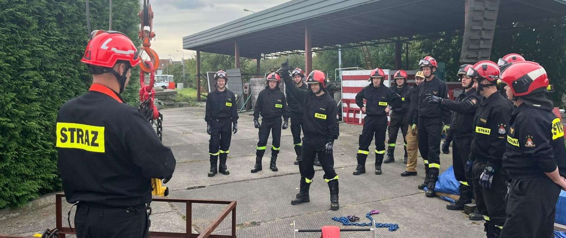 Strażacy Państwowej Straży Pożarnej podczas szkolenia "hakowego-sygnalisty". Jeden ze strażaków pokazuje znaki dla operatora żurawia aby podnosić na zawiesiach element stalowy. Pozostali strażacy w półkolu obserwują działania kolegi. Strażacy w ubraniach koszarowych z kaskami i hełmami na głowach. W tle widoczna wiata ze sprzętem na terenie Komendy Powiatowej PSP w Brzegu.