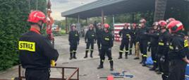 Strażacy Państwowej Straży Pożarnej podczas szkolenia "hakowego-sygnalisty". Jeden ze strażaków pokazuje znaki dla operatora żurawia aby podnosić na zawiesiach element stalowy. Pozostali strażacy w półkolu obserwują działania kolegi. Strażacy w ubraniach koszarowych z kaskami i hełmami na głowach. W tle widoczna wiata ze sprzętem na terenie Komendy Powiatowej PSP w Brzegu.