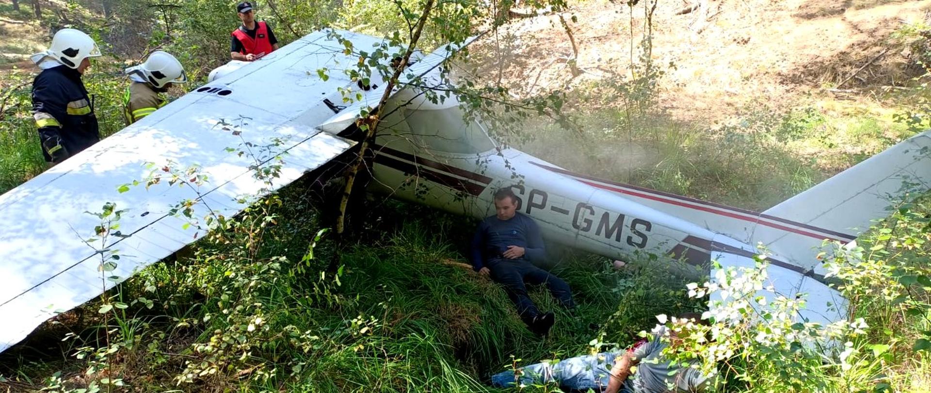 Zdjęcie zrobione na zewnątrz, podczas ćwiczeń. Mały samolot rozbity w lesie. Z samolotu wydobywa się dym. Obok stoi trzech strażaków.