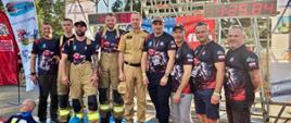 Grupowe zdjęcie ośmiu mężczyzn stojących na podium oznaczonym numerami 1, 2 i 3. Na najwyższym stopniu stoją strażacy w strojach sportowych i mundurach, w tym bryg. Jacek Cyburt - Komendant Miejski PSP w Szczecinie, w beżowym mundurze wyjściowym. Po ich bokach, na niższych stopniach i obok nich, stoją pozostali członkowie drużyny w koszulkach technicznych. U stóp podium umieszczone są strażackie rekwizyty: dwa fantomy szkoleniowe (ubrane w stroje ochronne) oraz czerwony i niebieski hydrant. W tle widać rusztowanie wieży ćwiczebnej, duży cyfrowy stoper wyświetlający czas oraz flagi sponsorów, m.in. "Panorama Morska". Nad podium wisi baner: "Mistrzostwa Województwa Zachodniopomorskiego o Tytuł Najtwardszego Strażaka, Jarosławiec 2026"