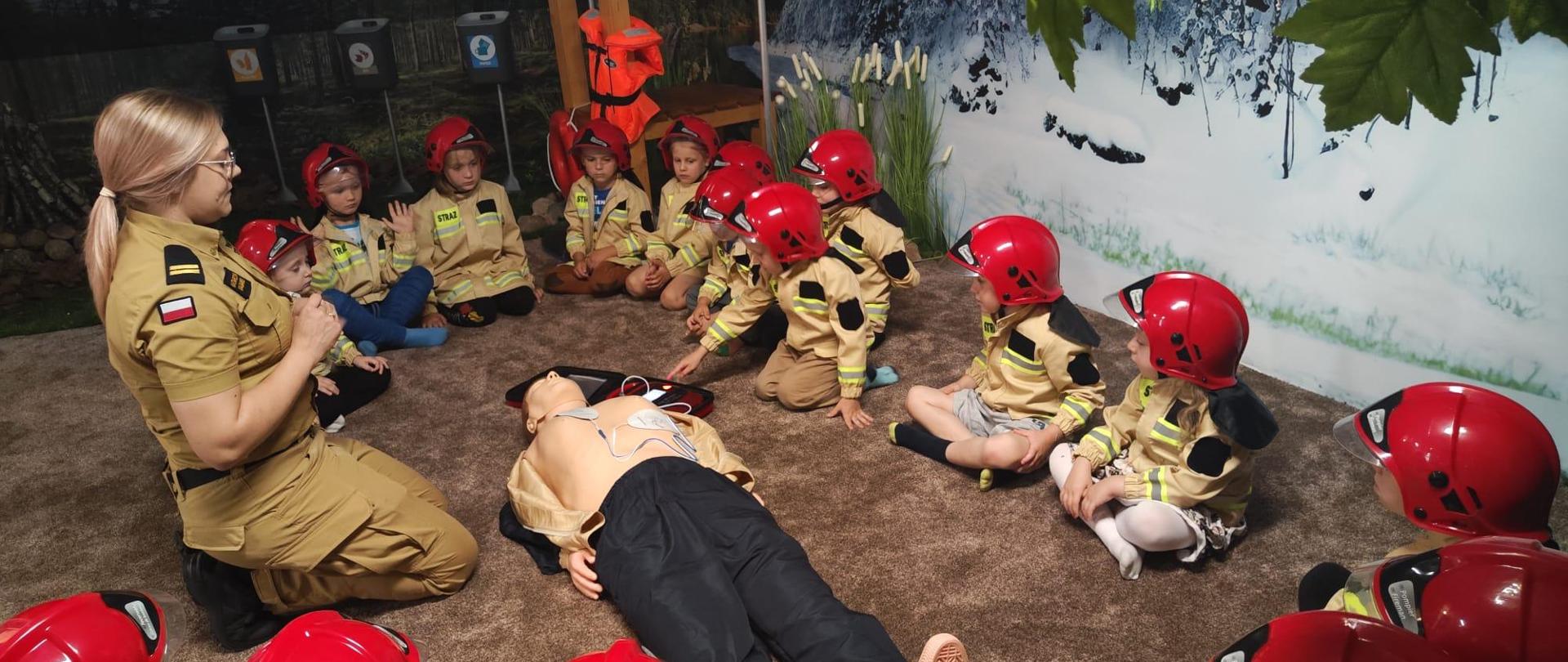 Dzieci w strojach strażaków słuchają kobiety strażak, która opowiada jak udzielać pierwszej pomocy