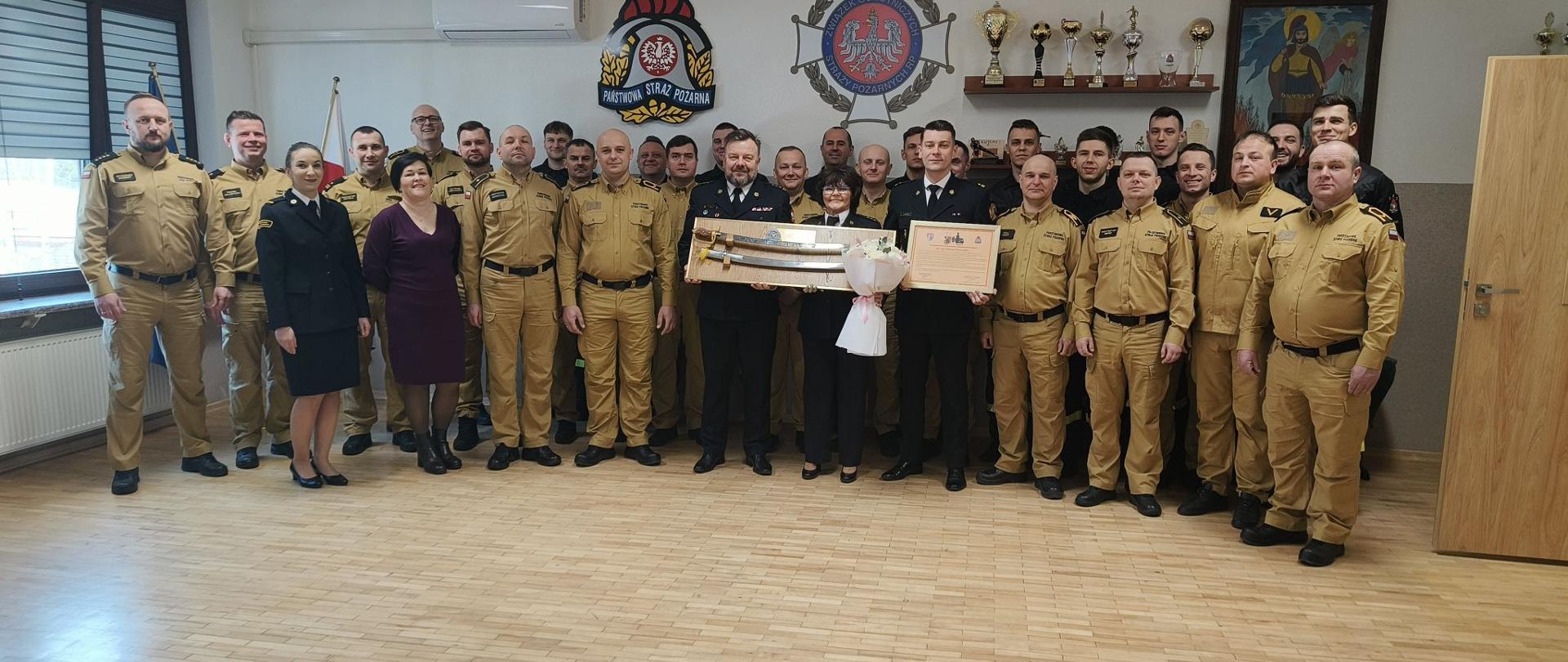 Zdjęcie przedstawia grupę kilkudziesięciu osób. Dwie z nich trzymają upominek, kobieta dodatkowo trzyma kwiaty, a mężczyzna obok trzyma list gratulacyjny.