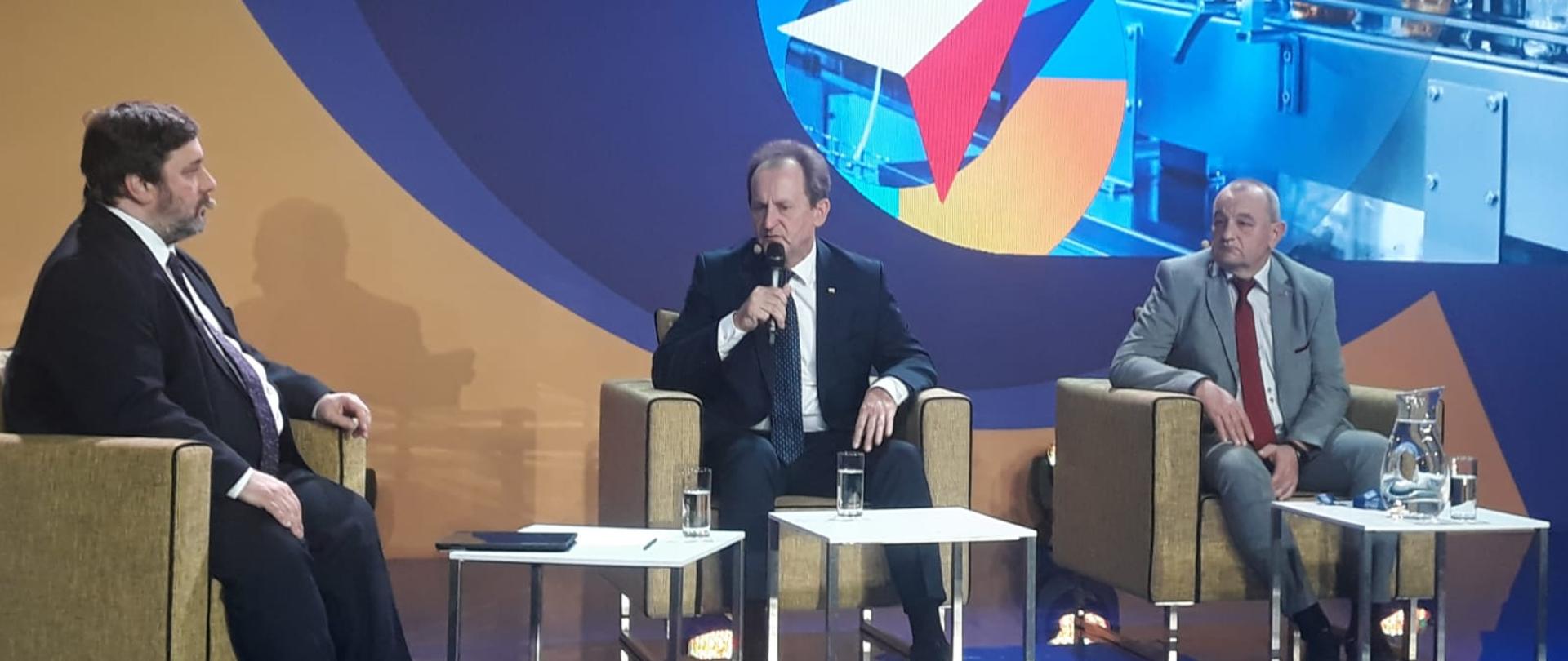 Na scenie siedzą trzej prelegenci. Przed nimi stoliki, na których stoją szklanki z wodą. Pomarańczowo-niebieskie to z trójkątną biało-czerwoną flagą. 