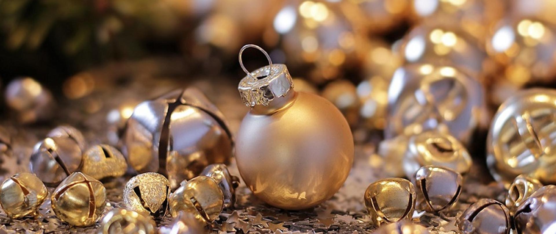 Na zdjęciu widać złote bombki i gwiazdki leżące pod choinką, w tle zielone gałązki choinki. 
