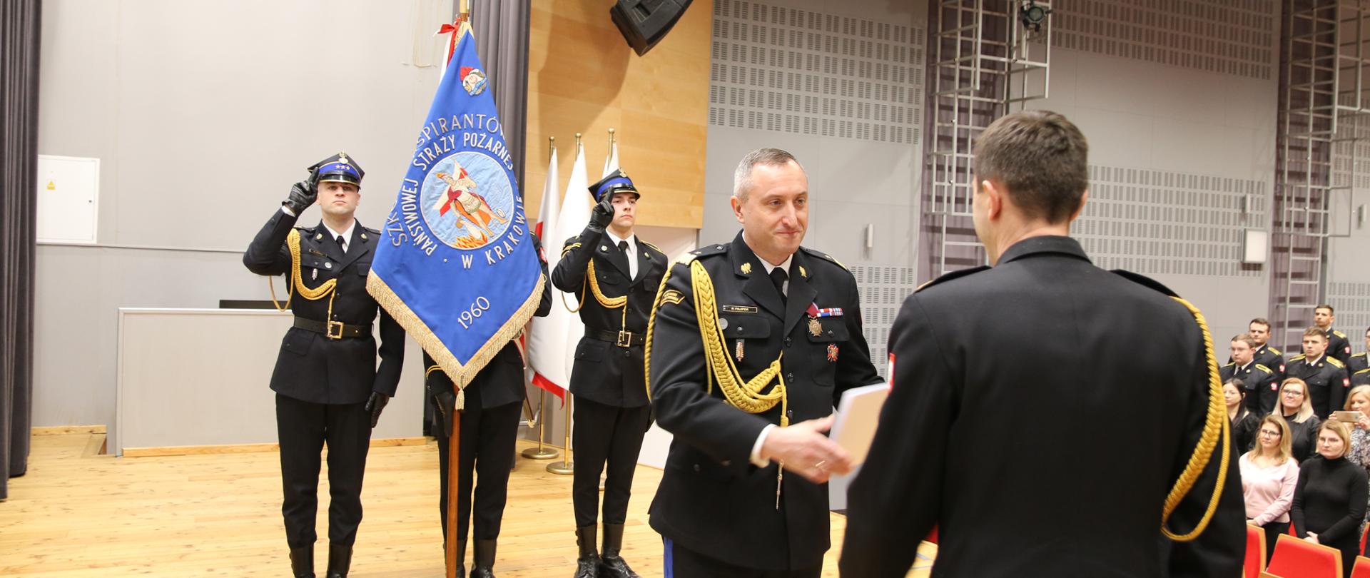 Nadbryg. Piotr Filipek całuje sztandar SA PSP w Krakowie odbiera decyzję personalną (białą teczkę) od stojącego tyłem Komendanta Głównego PSP, w tle sztandar Szkoły