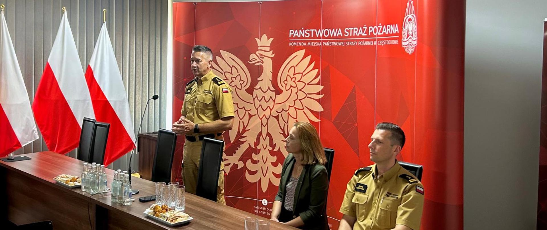 Funkcjonariusz Państwowej Straży Pożarnej w jasnobrązowym mundurze przemawia, stojąc za stołem prezydialnym. Obok niego siedzą kobieta w zielonej marynarce oraz drugi funkcjonariusz w mundurze. W tle znajduje się czerwona ścianka z dużym wizerunkiem orła oraz napisem „Państwowa Straż Pożarna – Komenda Miejska Państwowej Straży Pożarnej w Częstochowie”, a po lewej stoją polskie flagi.