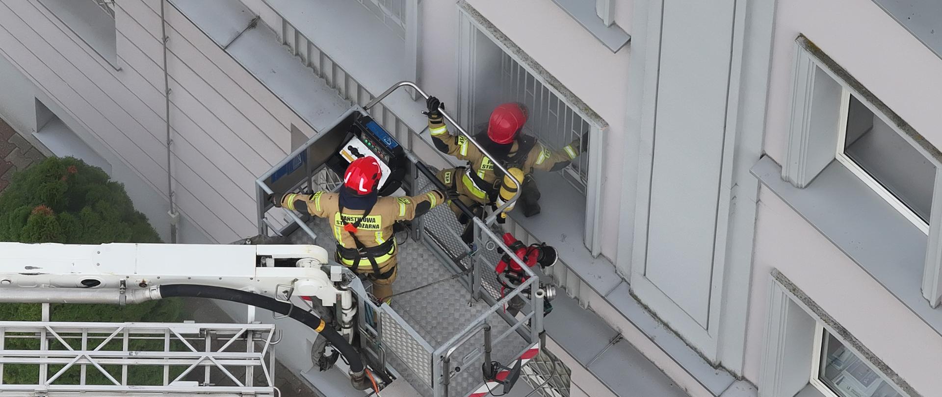 Strażacy na podnośniku zbliżają się do okna budynku. Z okna wydobywa się dym