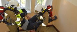Ćwiczenia w szpitalu. Dwóch strażaków w białych hełmach i piaskowych ubraniach ewakuuje osobę poszkodowaną. Jeden z nich przesuwa się tyłem trzymając ewakuowanego za nogi, drugi - przodem, trzymając za górne części ciała. Strażacy mają na plecach założony aparat powietrzny. W tle okna.