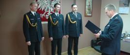 bryg. Robert Lebioda odczytuje decyzje o nadaniu stopnia kapitana dla mł. kpt. Grzegorza Olejarza oraz Dyplomu Komendanta Głównego PSP dla bryg. Krzysztofa Cebulaka Uczestnicy zbiórki w umundurowaniu wyjściowym. W tle godło polski. 