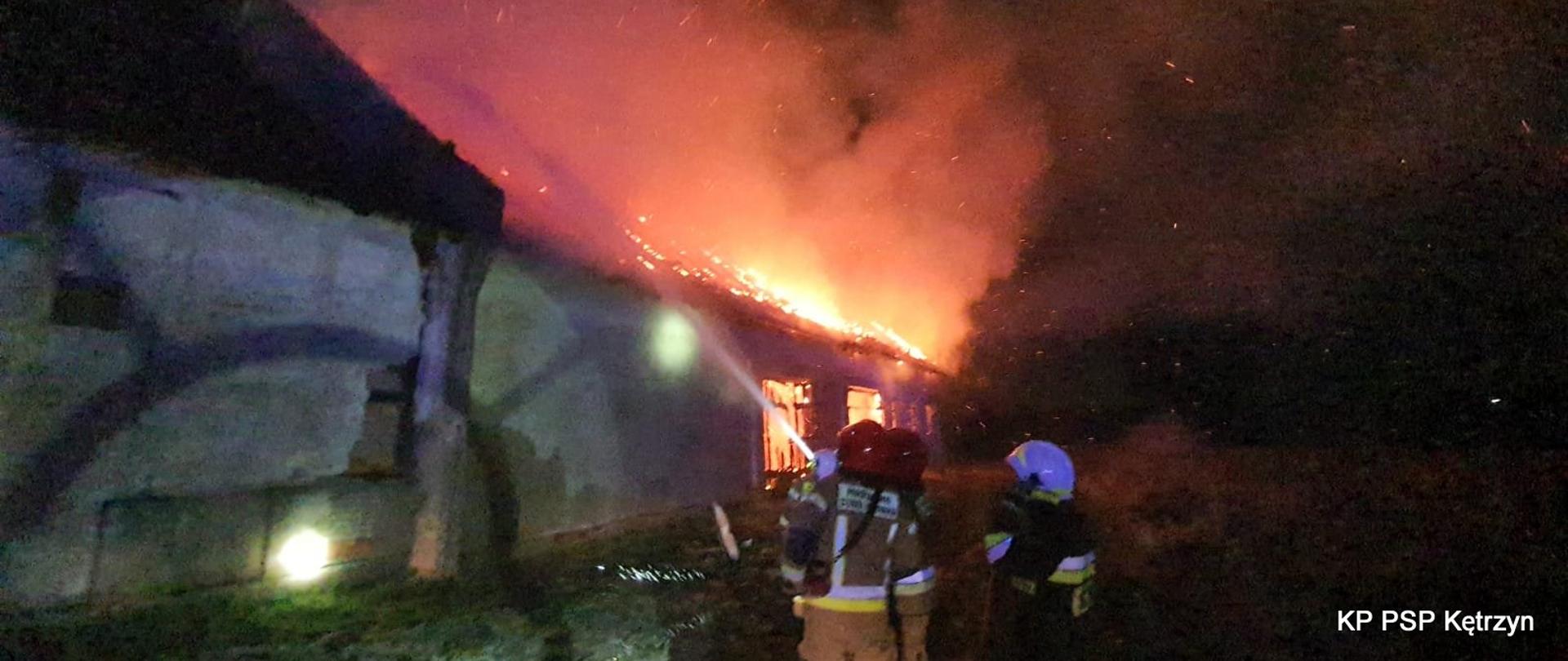 Zdjęcie wykonane na miejscu zdarzenia podczas nocnego pożaru stodoły. Strażacy stoją przed budynkiem i leją wodę na budynek objęty pożarem. Biały bym unosi się nad budynkiem wraz z ogniami lotnymi.