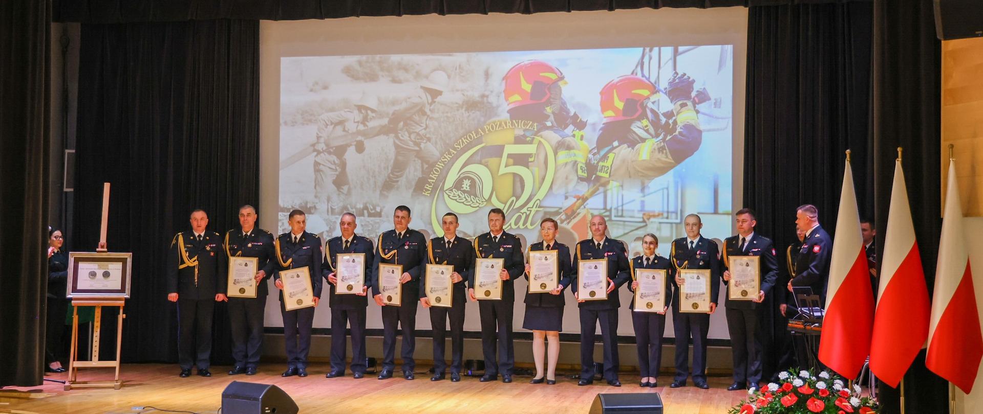 ALT: Na scenie stoi grupa odznaczonych funkcjonariuszy i pracowników służb, trzymających pamiątkowe grawertony. W tle widoczna jest projekcja z logo 65-lecia szkoły pożarniczej.