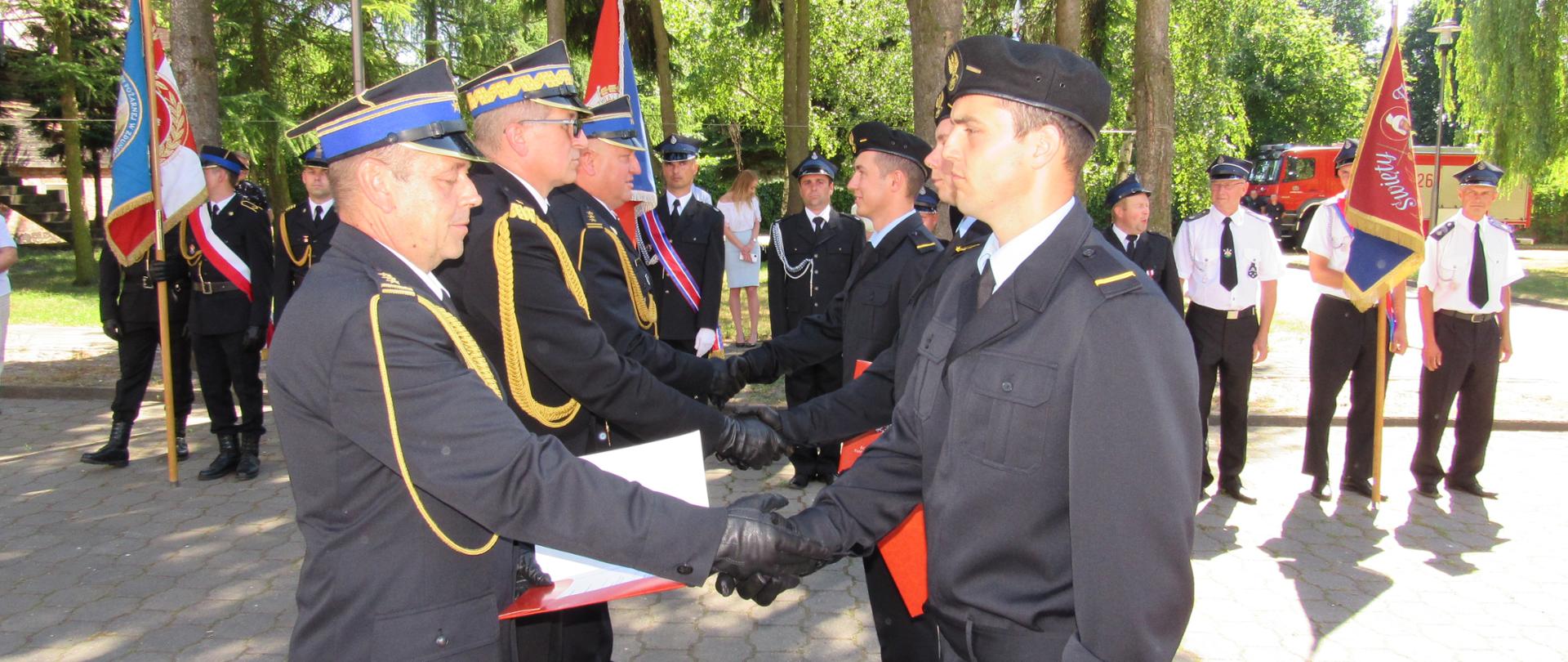 Na zdjęciu widać komendanta powiatowego PSP w zduńskiej woli gratulującego awansu strażakowi za nim stoją komendant wojewódzki PSP w łodzi, oraz zastępcę komendanta głównego PSP, w tle poczty sztandarowe, i uczestnicy powiatowych obchodów dnia strażaka