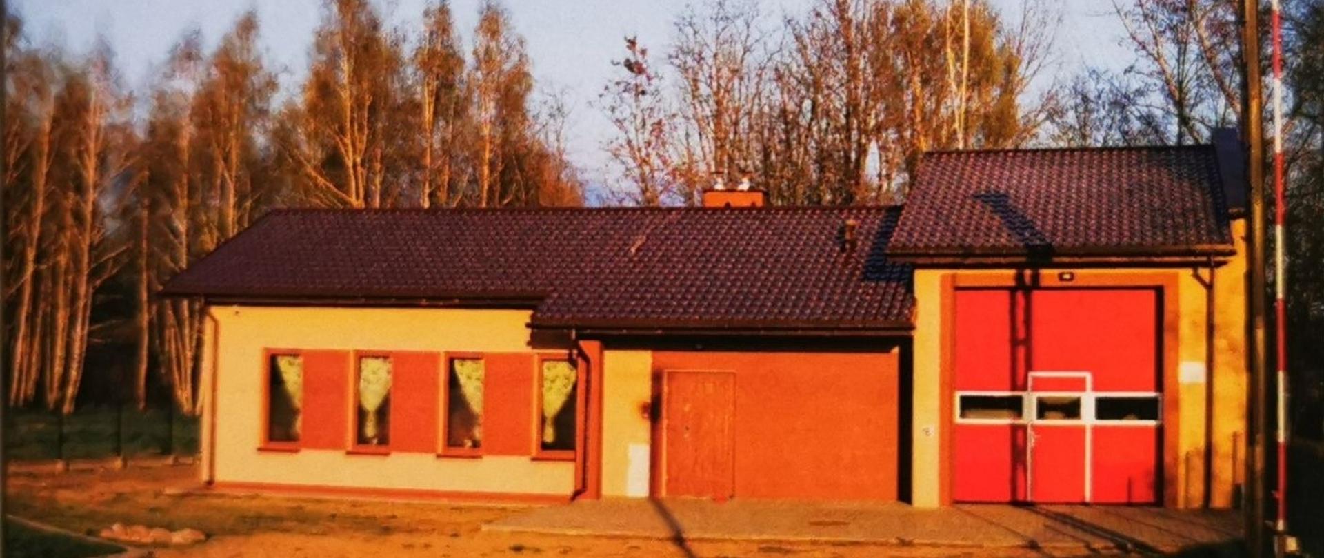 Zdjęcie przedstawia obecny widok na remizę OSP Borki Prusinowskie. Z prawej strony duża czerwona brama garażowa na samochód OSP z lewej strony wejście na świetlicę oraz 4 okna. Budynek pokryty blachodachówką. W tle widoczne drzewa. 