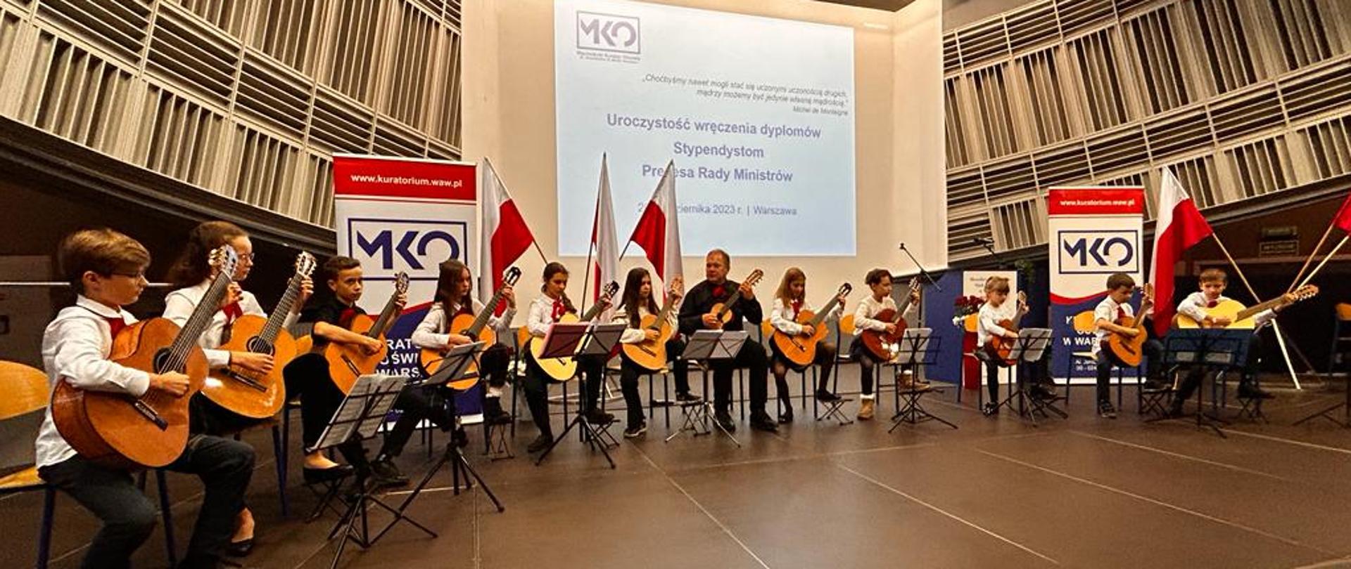 Na zdjęciu widać cały zespół siedzący w półkolu, wykonujący utwór, w tle rzutnik ze slajdem tytułowym