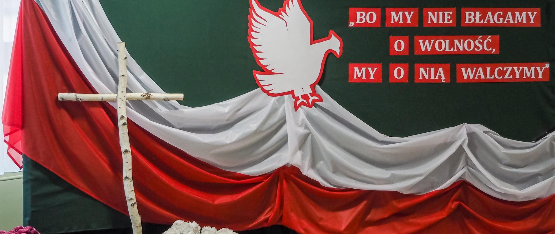 Na fotografii patriotyczna scenografia z napisem „Bo my nie błagamy o wolność, my o nią walczymy”, orzeł z biało- czerwoną, krzyż brzozowy i białe chryzantemy.