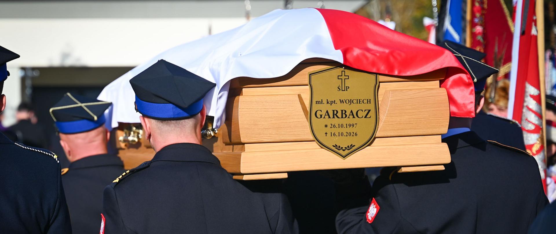 Uczestnicy ceremonii pogrzebowej na cmentarzu, wśród nich przedstawiciele kierownictwa Państwowej Straży Pożarnej. Na pierwszym planie widoczny wieniec z biało-czerwonymi kwiatami.