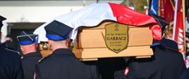 Uczestnicy ceremonii pogrzebowej na cmentarzu, wśród nich przedstawiciele kierownictwa Państwowej Straży Pożarnej. Na pierwszym planie widoczny wieniec z biało-czerwonymi kwiatami.