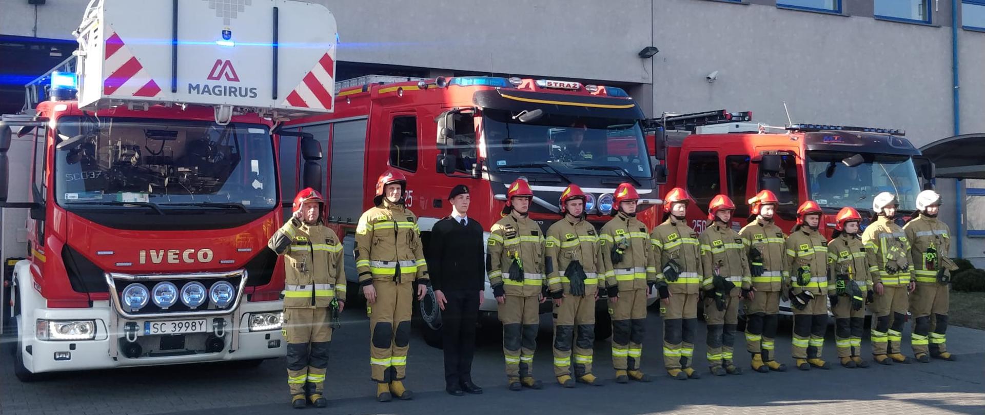 Strażacy stoją w szeregu przed garażami Jednostki Ratowniczo-Gaśniczej. Za nimi wystawione wozy bojowe z włączonymi sygnałami świetlnymi