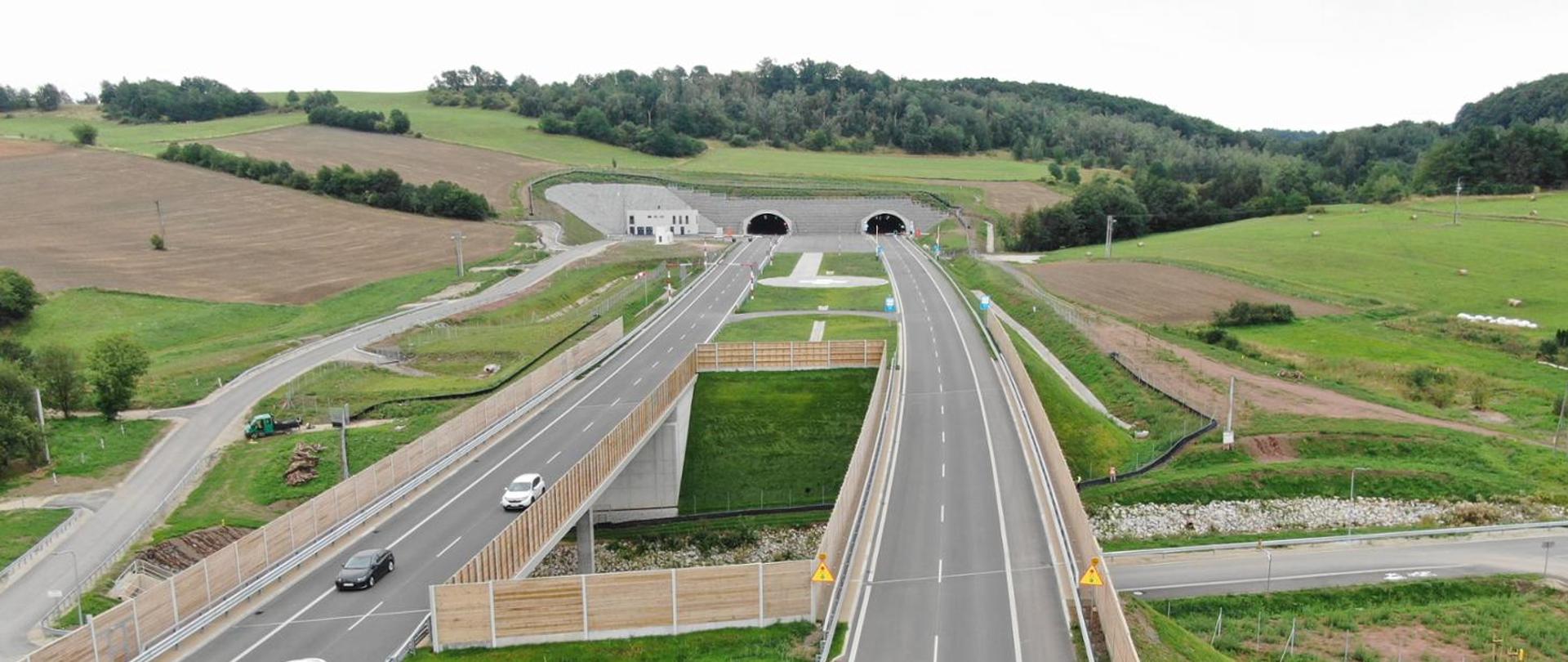 Widok z lotu ptaka na drogę ekspresową S3 prowadzącą do dwunawowego tunelu drogowego. Przed wjazdem do tunelu jezdnie rozdzielają się i przebiegają przez zielony, pagórkowaty krajobraz z polami uprawnymi i lasem w tle. Na drodze widocznych jest kilka samochodów.