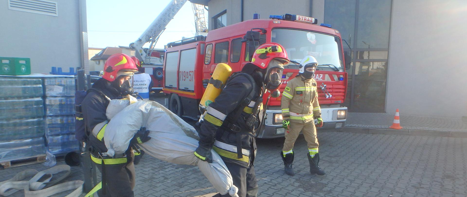 Przed budynkiem biedronki dwóch strażaków w umundurowaniu specjalnym wynosi manekina. Za nimi widać trzeciego strażaka, który idzie i samochód ratowniczo- gaśniczy.
