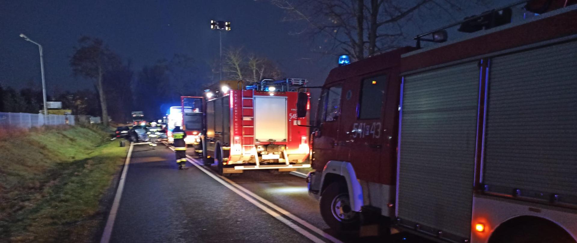 Na zdjęciu samochody strażackie stojące na drodze. W dalszym planie widać jeden z uszkodzonych samochodów, który znajduje się na jezdni. Zdjęcie wykonane w godzinach porannych.