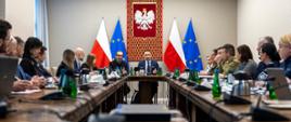 Spotkanie rządowe w sali konferencyjnej. Uczestnicy siedzą przy stole w kształcie podkowy. Na czele stołu przemawia mężczyzna na tle flag Polski i UE. Obecni są także przedstawiciele wojska i administracji.