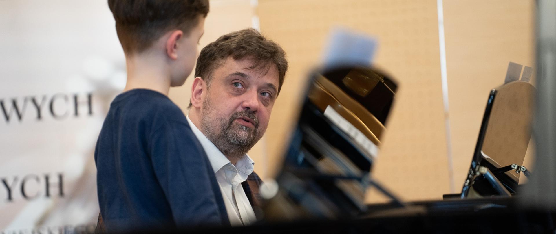 zdjęcie przedstawia prowadzącego warsztaty w rozmowie z uczestnikiem warsztatów przy dwóch fortepianach na scenie sali koncertowej, ujęcie z bliska