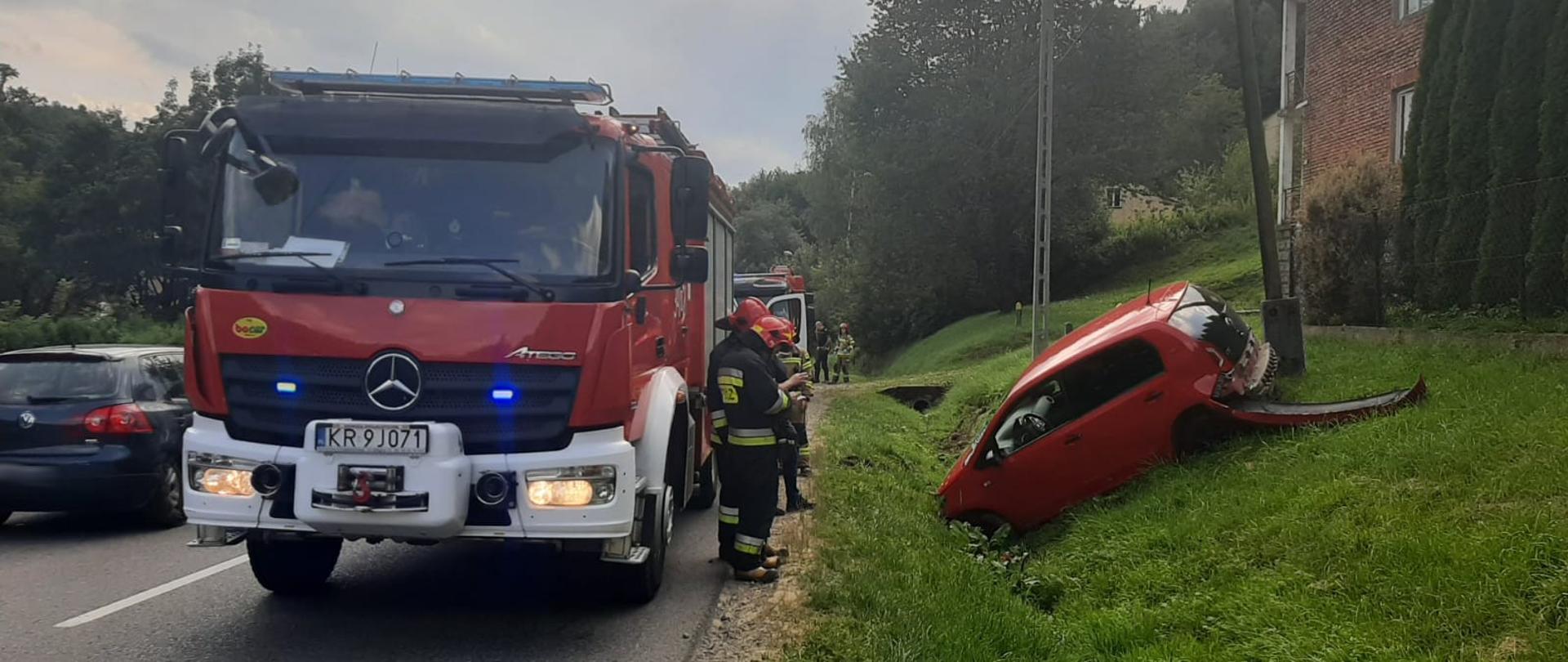 Samochód pożarniczy na drodze. Obok niego strażacy a poza drogą samochód osobowy.