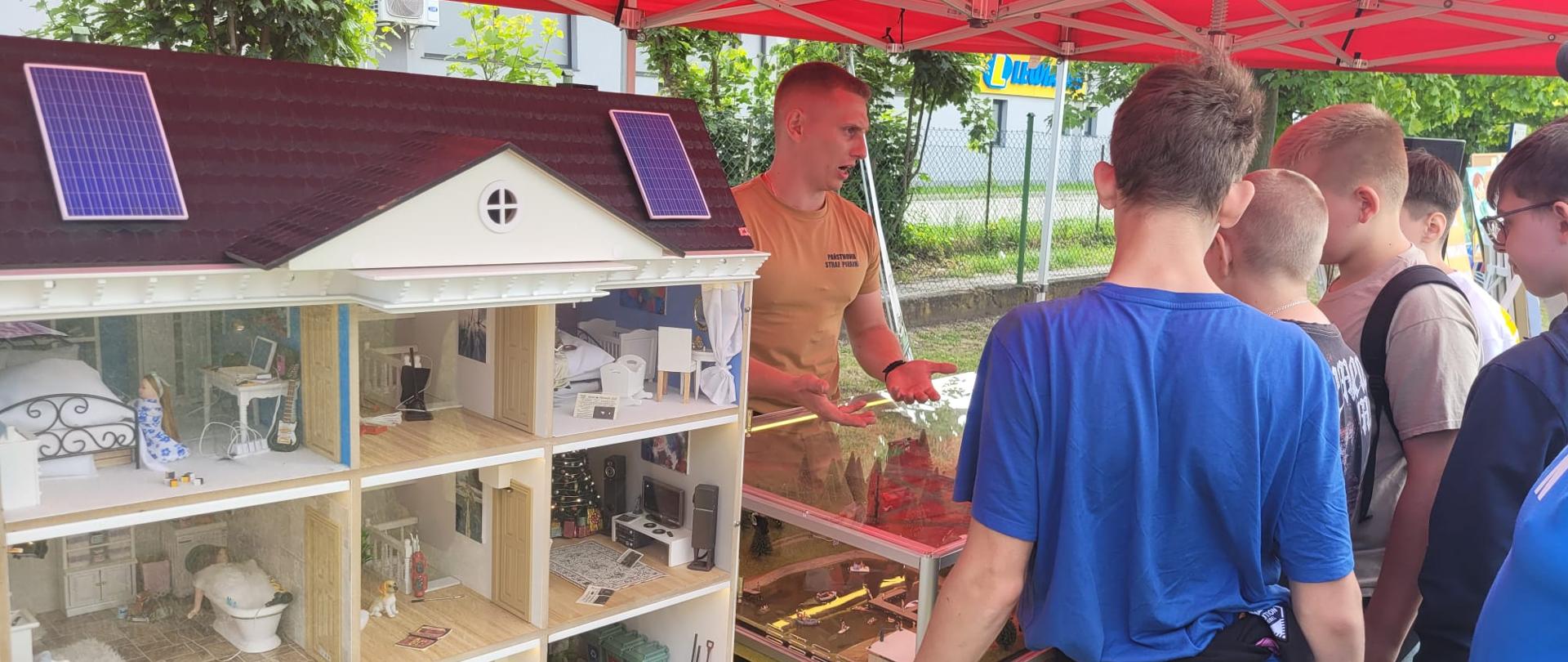 Pod czerwonym namiotem Komendy Powiatowej Państwowej Straży Pożarnej w Kole odbywa się pokaz edukacyjny. Strażak tłumaczy grupie młodzieży zasadę działania interaktywnego modelu domu, który przedstawia różne pomieszczenia wyposażone w miniaturowe meble i elementy codziennego użytku. Na dachu modelu znajdują się panele fotowoltaiczne. Młodzież z zaciekawieniem słucha prezentacji, a strażak wskazuje ręką na elementy ekspozycji. Celem pokazu jest edukacja w zakresie zagrożeń pożarowych i bezpiecznego zachowania w domu.