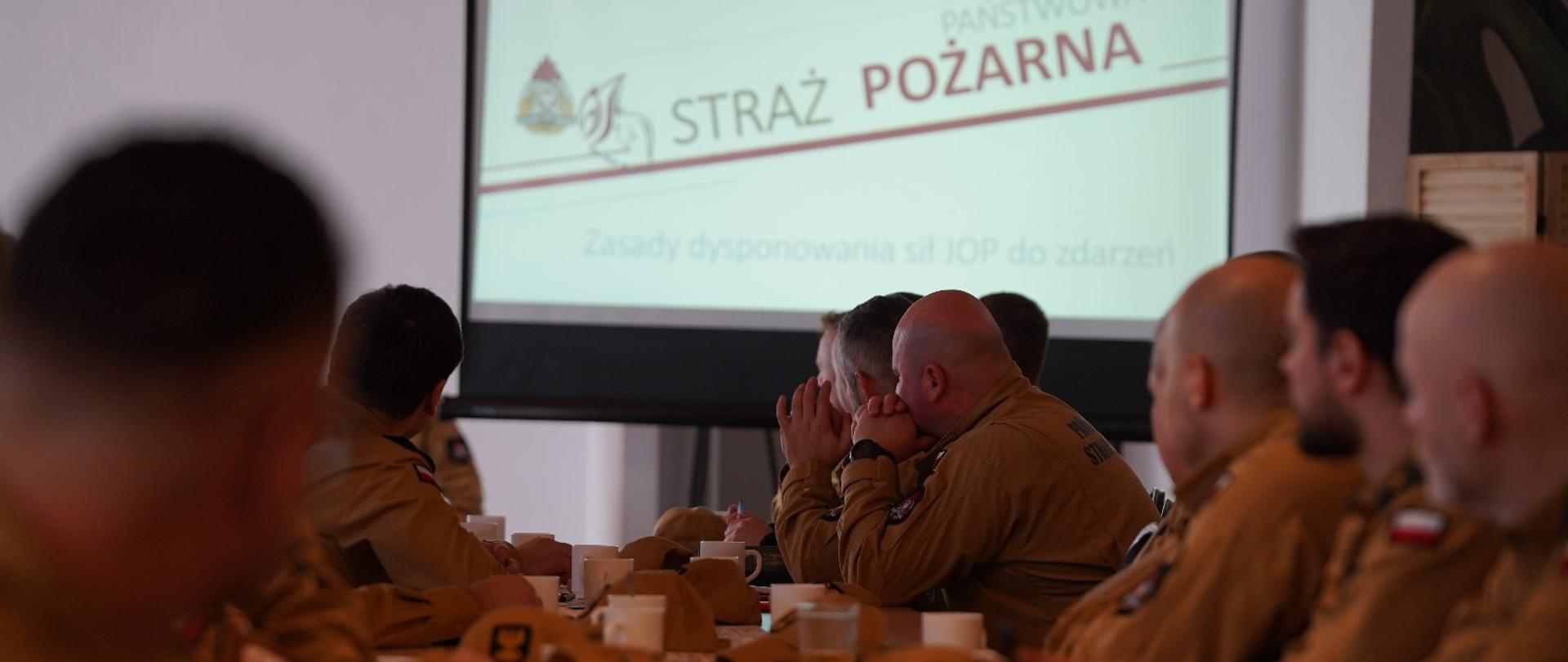 Na zdjęciu widzimy grupę strażaków Państwowej Straży Pożarnej uczestniczących w spotkaniu lub szkoleniu. Wszyscy mają na sobie charakterystyczne mundury w kolorze żółto-brązowym z naszywkami polskiej flagi oraz napisem „PAŃSTWOWA STRAŻ POŻARNA” na plecach. Strażacy siedzą przy długich stołach, na których znajdują się filiżanki z napojami i dokumenty.
Na pierwszym planie kilku strażaków siedzi tyłem do obiektywu, zwróconych w stronę prelegenta, który stoi przy stole i przemawia. W tle widoczny jest ekran z napisem „STRAŻ POŻARNA” oraz logo, co sugeruje, że trwa oficjalna prezentacja. Sala jest przestronna, ma drewniane elementy wystroju oraz zielone rośliny na ścianach, co nadaje jej przyjazny charakter.
Cała scena wygląda na formalne spotkanie lub naradę, gdzie strażacy omawiają ważne kwestie związane z ich służbą.