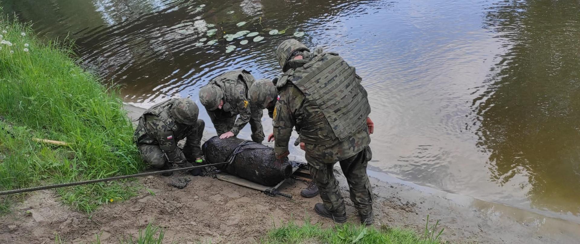 Na brzegu rzeki stoi czterech żołnierzy w zielonych mundurach. Na piasku leży przedmiot przypominający bombę.