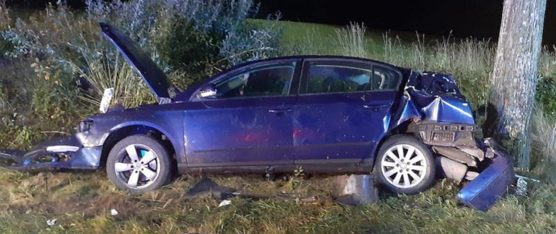 Zdjęcie przestawia rozbity pojazd koloru niebieskiego, który w wyniku zderzenia z drzewem znajduje się na poboczu drogi. Auto ma widoczne uszkodzenia zarówno z przodu jak i z tyłu. Wkoło leżą porozrzucane elementy karoserii.