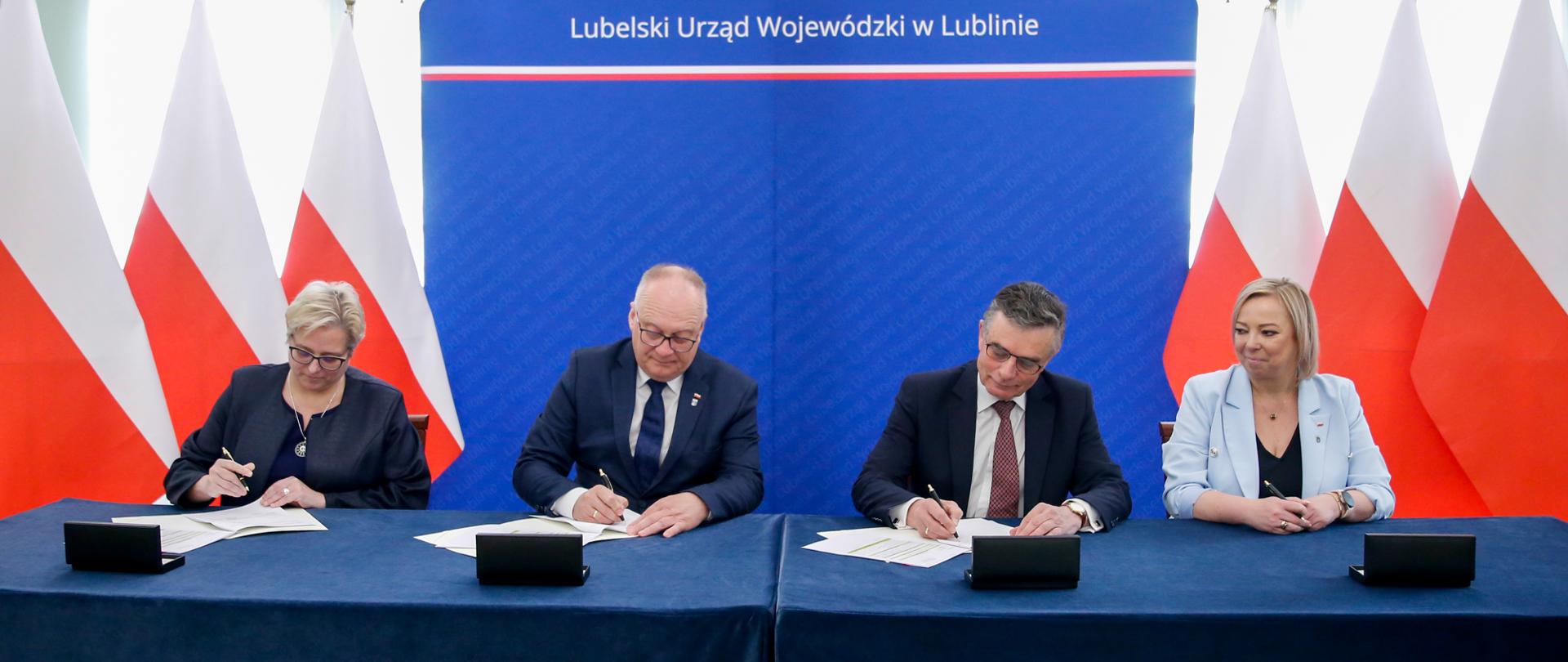 Dwóch mężczyzn i dwie kobiety siedzą przy stole prezydialnym. Podpisują umowy.