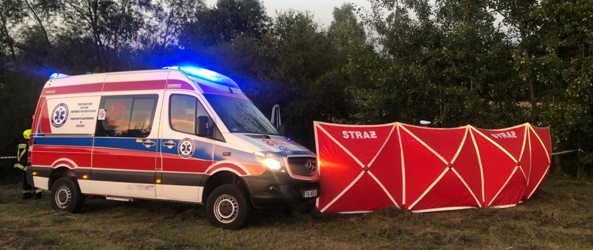 Zdjęcie przedstawia miejsce zdarzenia. Po lewej stronie stoi ambulans Państwowego Ratownictwa Medycznego tuż przy rozstawionym strażackim parawanie osłaniającym.