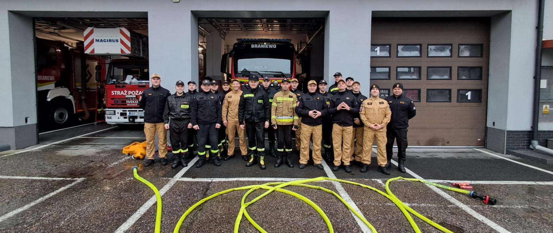 Zdjęcie grupowe strażaków przy remizie jednostki. W tle samochody strażackie w garażu.