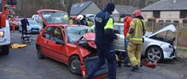 Na jezdni i pobpoczu drogi stoja dwa rozbite pojazdy osobowe. Jeden koloru srebrnego, drugi - czerwonego. Pomiędzy nimi strażacy i policjanci prowadzą akcję ratowniczą. Za nimi pojazdy strażackie i policyjne. W tle zabudowania i las. 