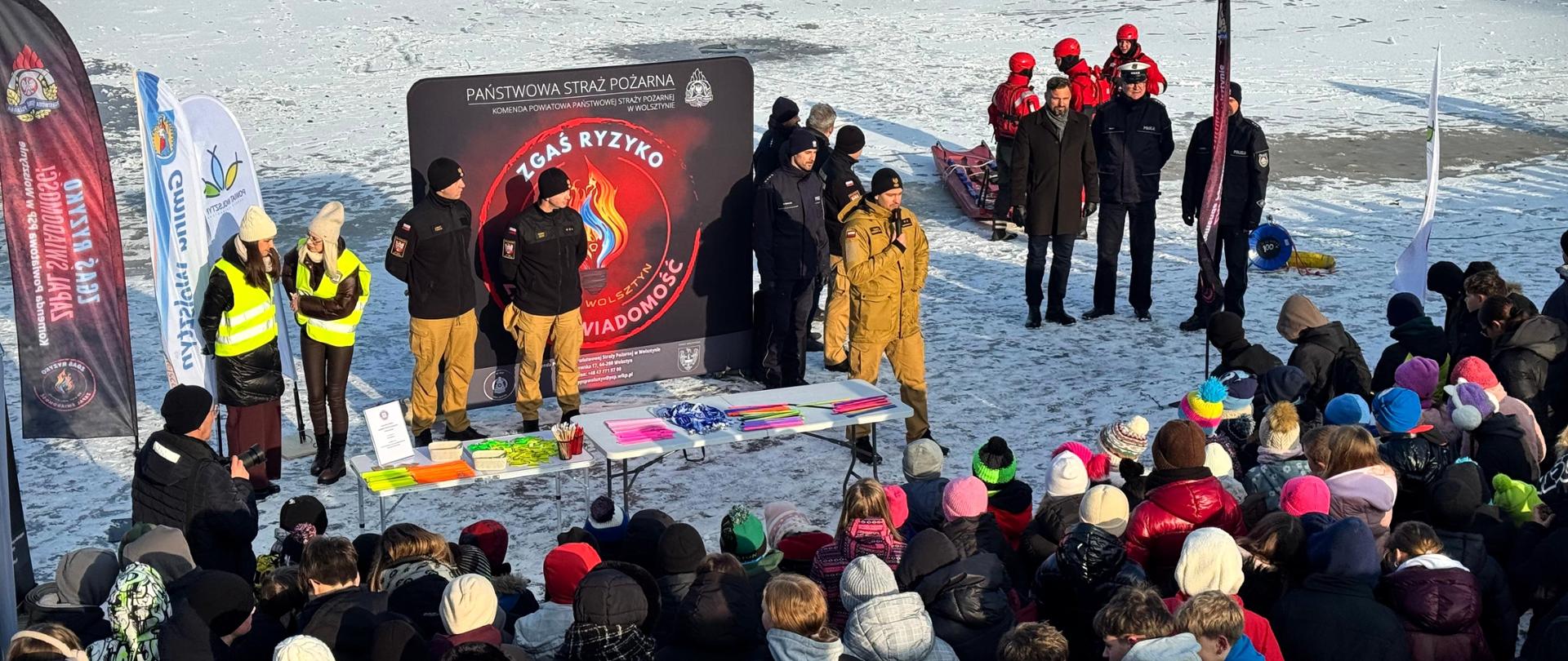 Zdjęcie przedstawia grupę funkcjonariuszy i przedstawicieli służb stojących w jednym szeregu przy zamarzniętym jeziorze. Wśród nich widoczni są strażacy Państwowej Straży Pożarnej w umundurowaniu służbowym, policjanci oraz przedstawiciele innych służb. Strażak trzyma mikrofon i przemawia do zgromadzonych uczestników. W tle znajdują się banery informacyjne Państwowej Straży Pożarnej z hasłem „Zgaś ryzyko zapal świadomość”. Zdjęcie ukazuje oficjalne rozpoczęcie wydarzenia plenerowego.